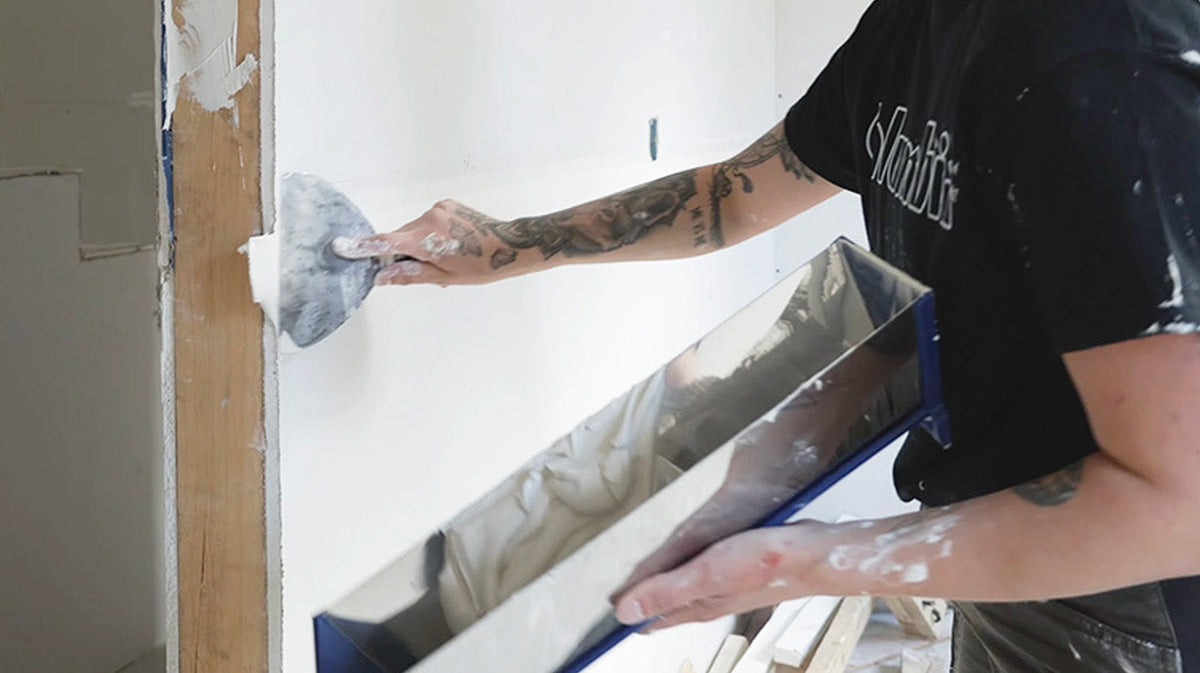 Drywaller applying joint compound over drywall tape with a joint knife while holding a mud pan